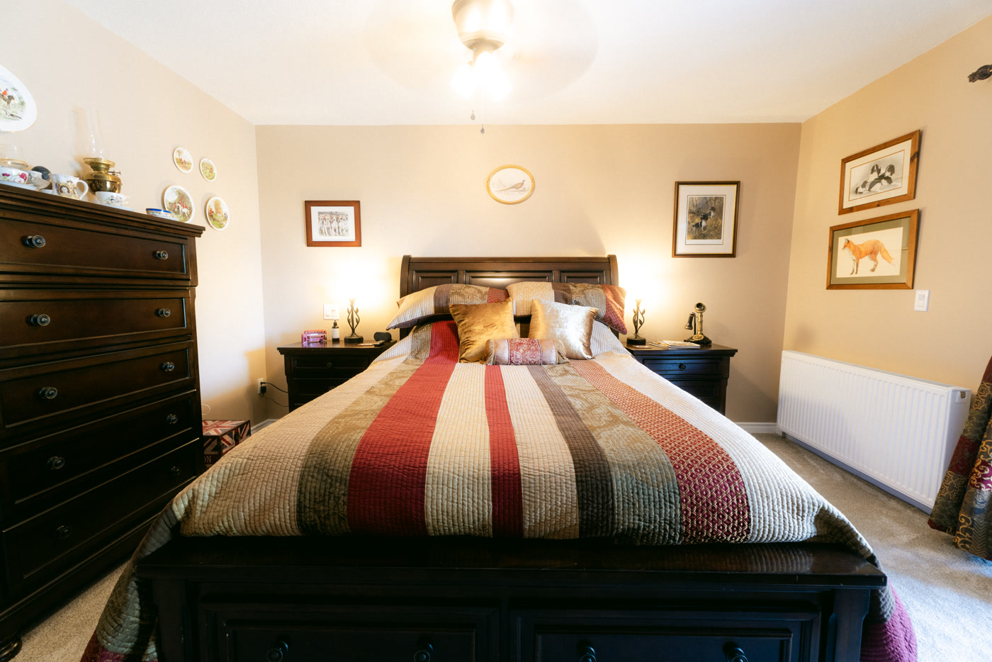 The Windsor Room's queen bed head-on at Shire Ridge Lodge near Bragg Creek: a quilt striped in red, cream and brown, lamps on both nightstands, a tall dark dresser at the side