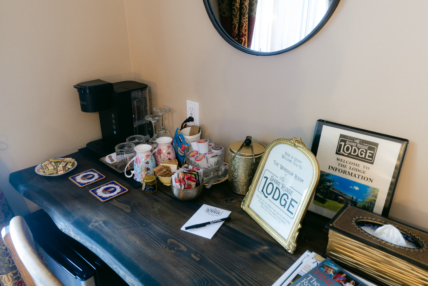 The desk in the Windsor Room at Shire Ridge Lodge near Bragg Creek: a coffee machine, mugs, a tray of treats, a framed lodge welcome sign and a round mirror above