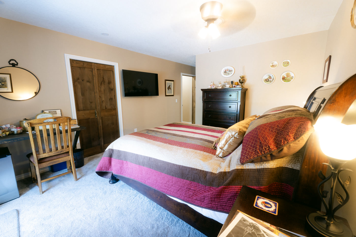 The Windsor Room from its corner at Shire Ridge Lodge near Bragg Creek: the striped bed, a wooden chair at the desk, a television, a dark dresser and plates on the wall