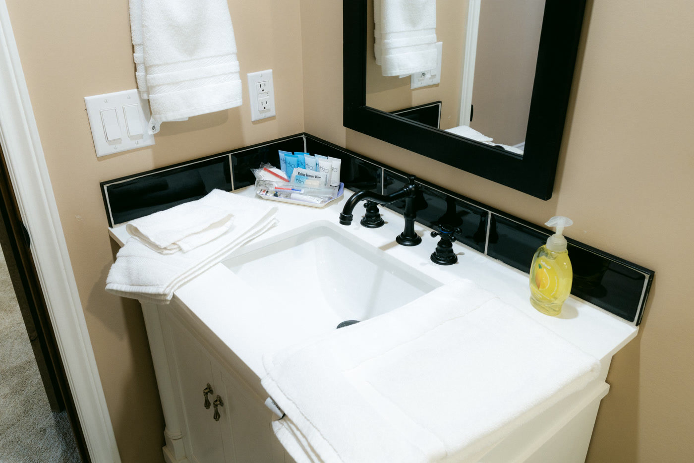 The Windsor Room's white vanity at Shire Ridge Lodge near Bragg Creek: a black faucet, folded white towels, a soap dispenser, a black tile backsplash and a dark framed mirror