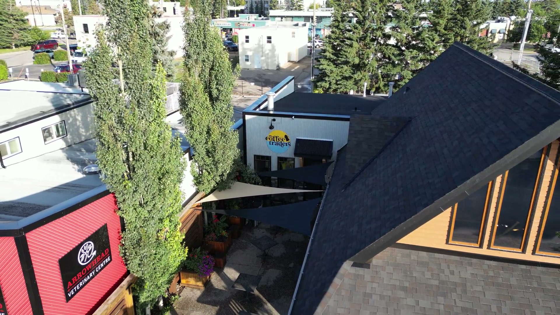Pulling back and rising from the round yellow-and-blue sign in the courtyard of Cochrane Coffee Traders, over shade sails and tall poplars, until the red clinic next door and the timber gable come into frame