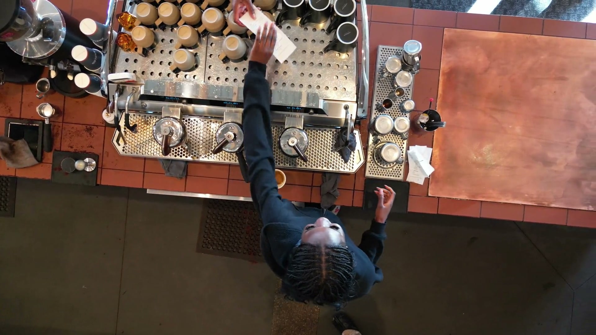Straight down over the espresso bar from ceiling height, two baristas working the machine, rows of cups on its steel top