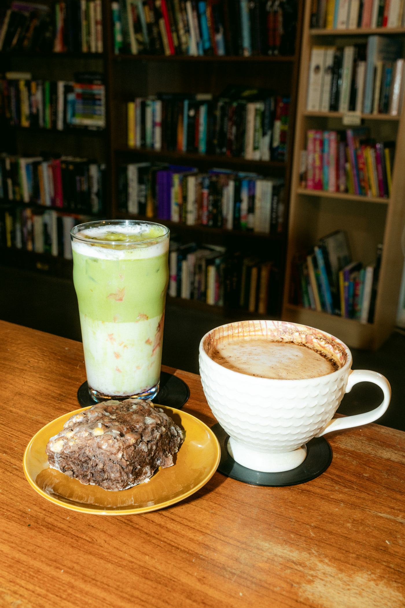 A tall glass of pale green matcha in the foreground at Found Books & Shop in Cochrane, the packed bookshelves standing behind it