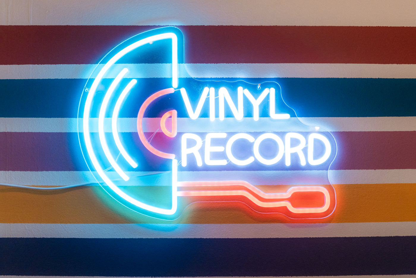 A neon sign reading VINYL RECORD on a wall painted in blue, white, red and orange stripes in the record room at Found Books & Shop in Cochrane