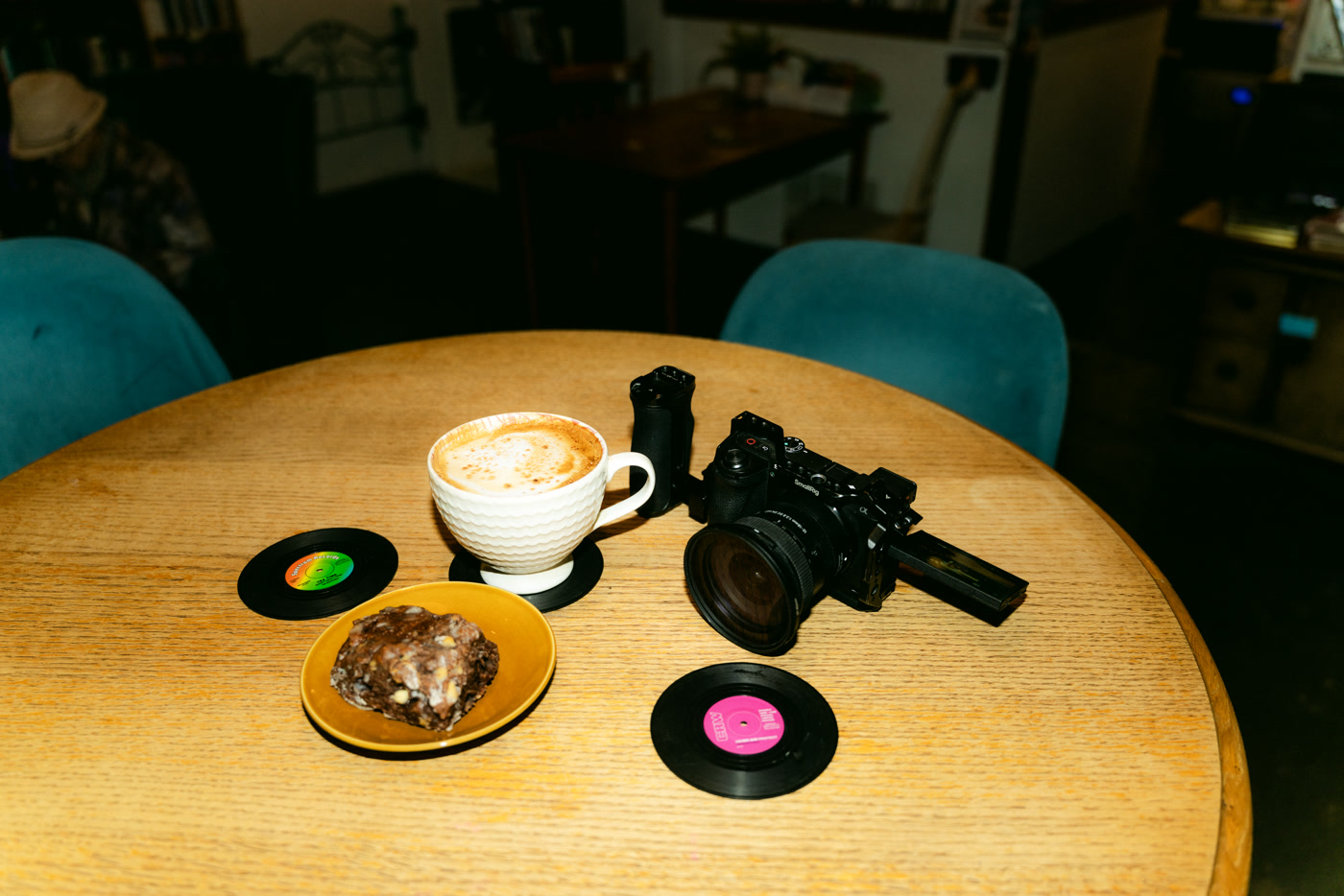 A round pale-wood table in the café at Found Books & Shop in Cochrane: a cappuccino, a pastry on a yellow plate, a camera set down on the wood and two seven-inch singles used as coasters