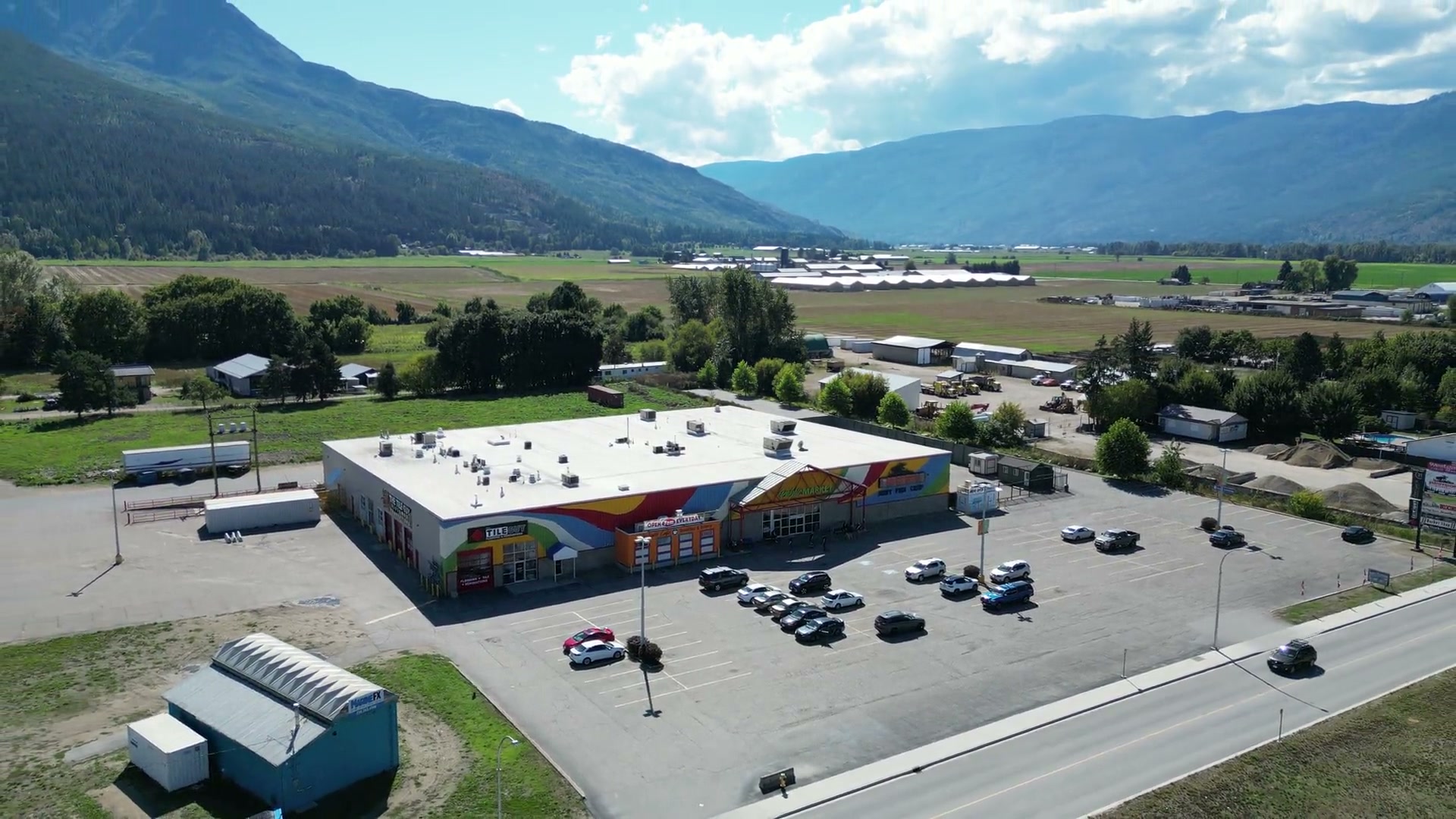 From high over the lot, pulling back and up until Westgate Public Market is one small white roof in the valley at Salmon Arm, fields, a road and the mountains all around it