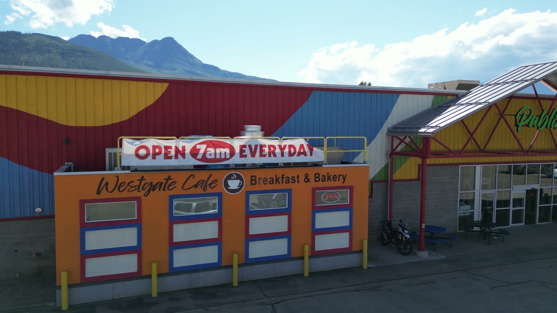 Sliding right along the orange front of Westgate Cafe in Salmon Arm, past its four red-and-blue windows and the lit OPEN sign, to the red steel gable over the market's door