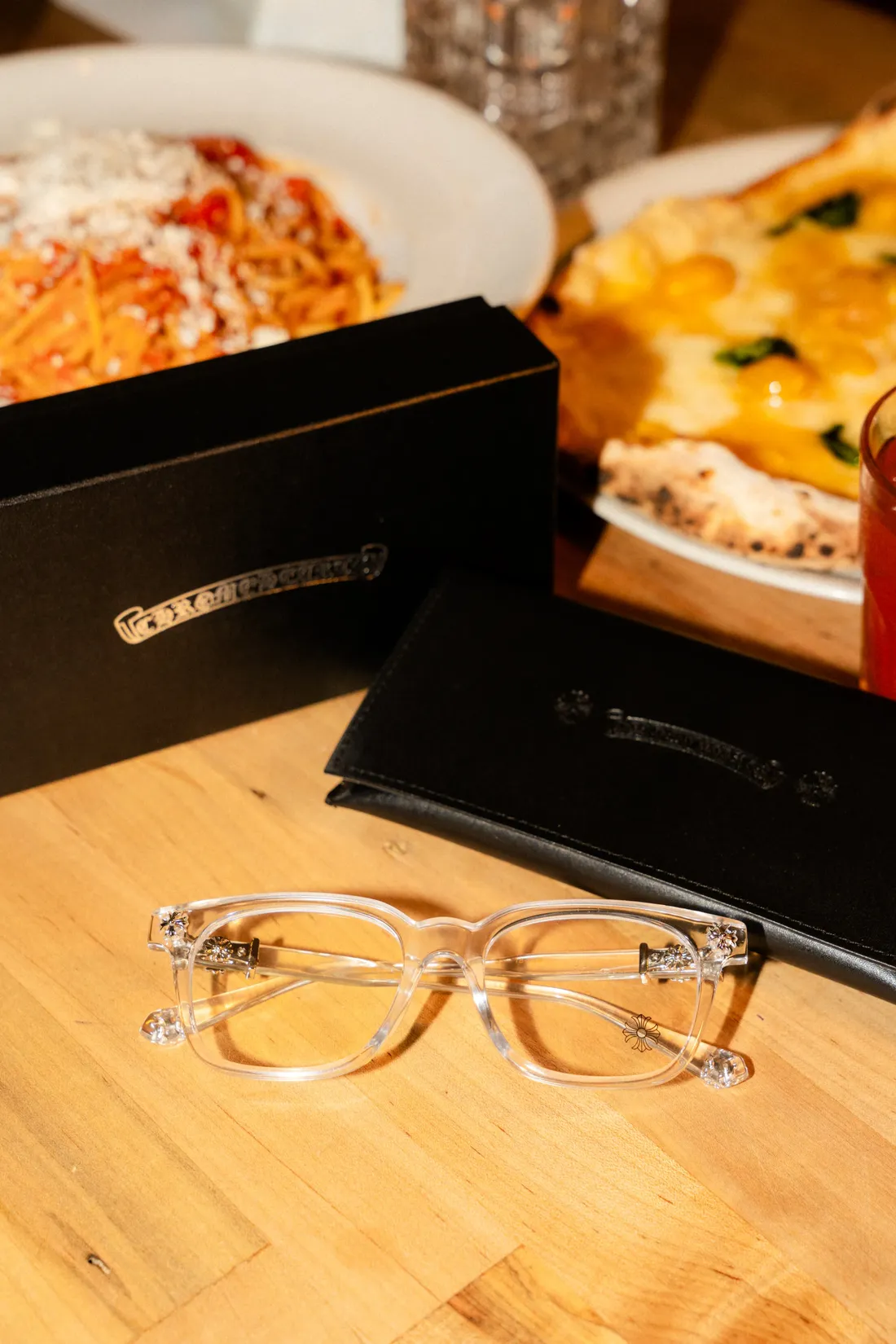 Clear eyeglasses and their case on a warm wooden table set with pasta and pizza