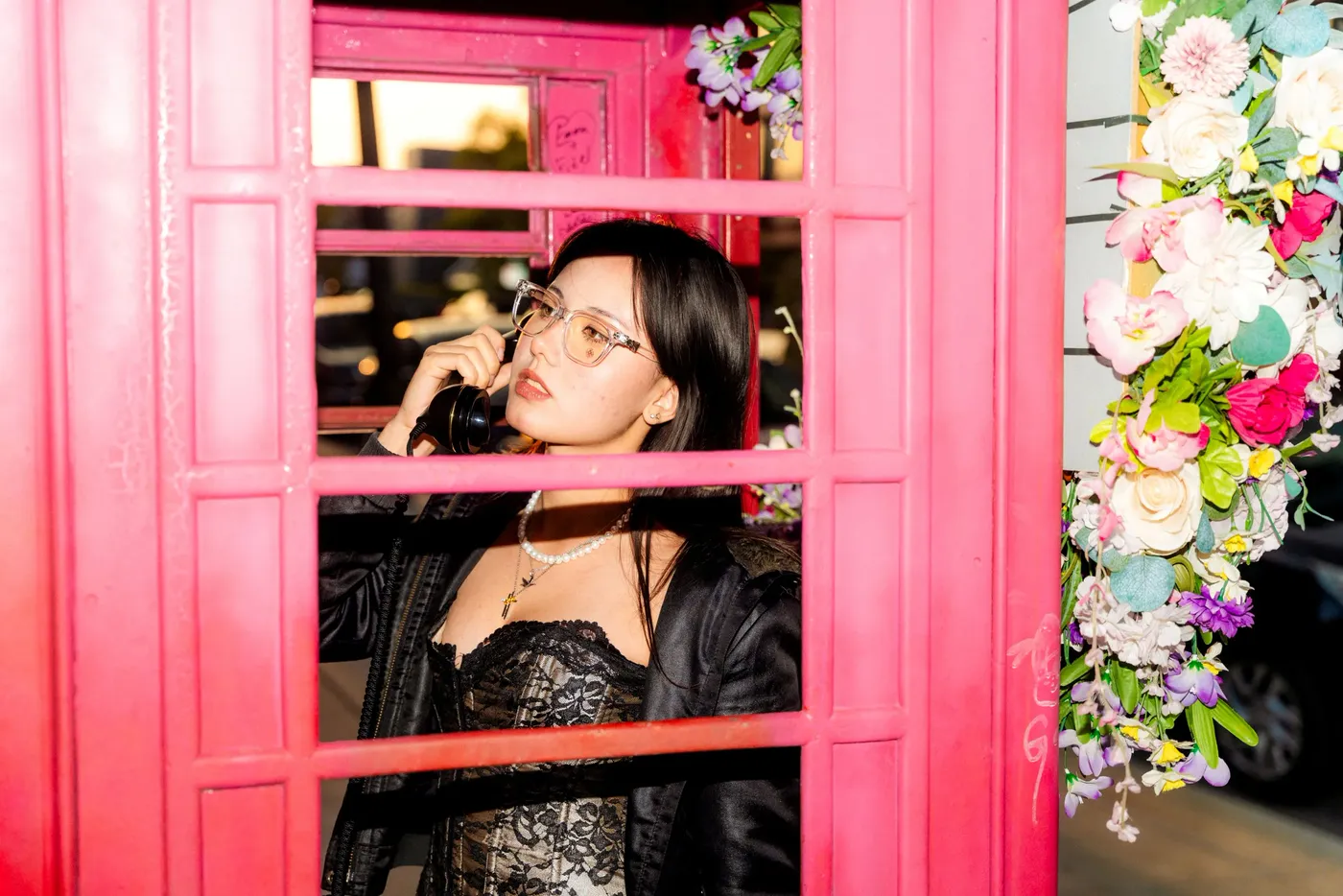 A woman in eyeglasses and a leather jacket leaning inside a bright pink phone booth trimmed with flowers