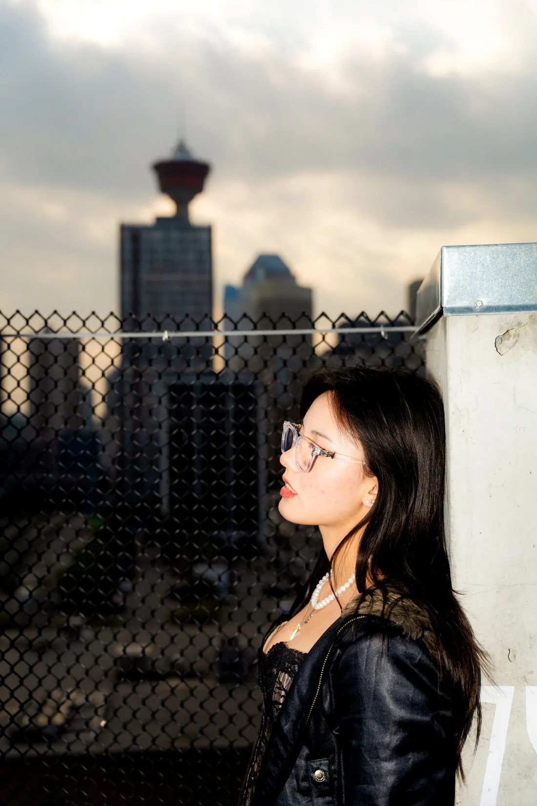 A woman in eyeglasses on a rooftop, the city skyline behind a chain-link fence