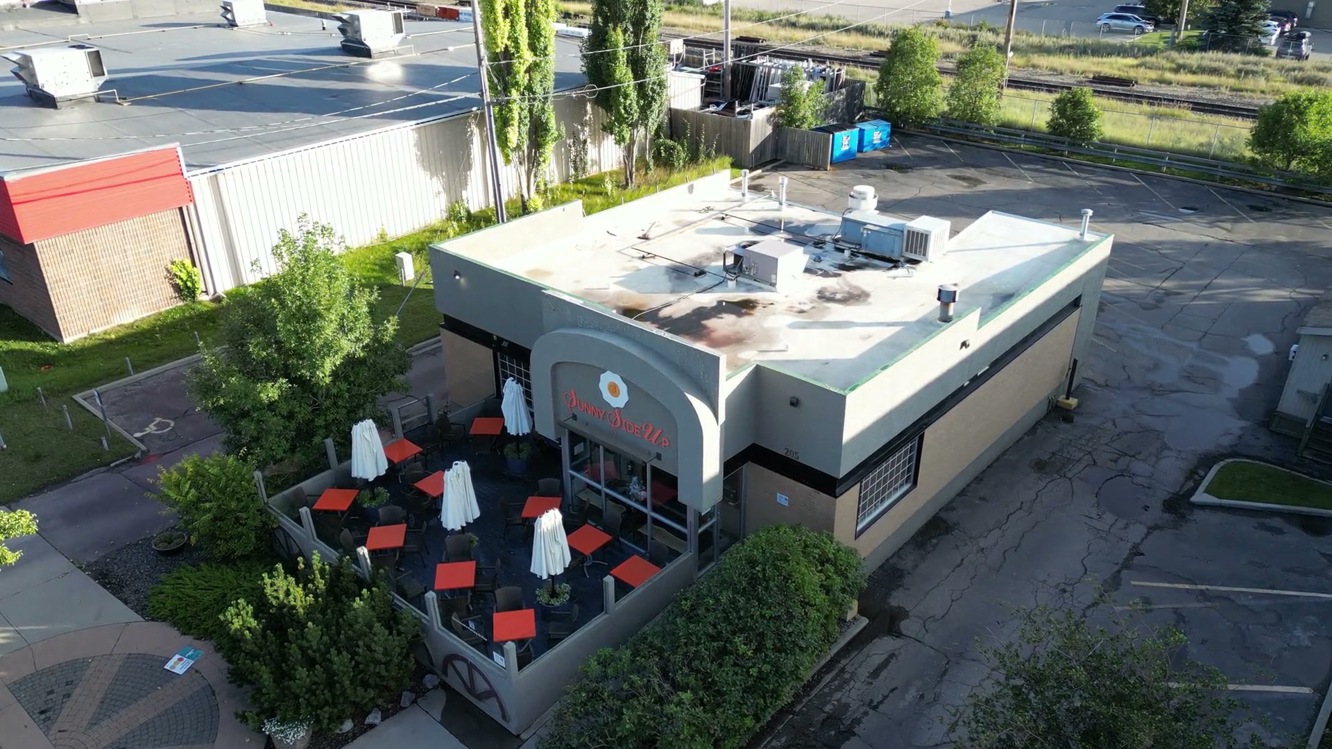 Hanging well above the corner of Sunny Side Up in Cochrane, the camera barely drifting: the walled patio of red tables and furled white umbrellas, the arched sign, the flat roof, the parking lot