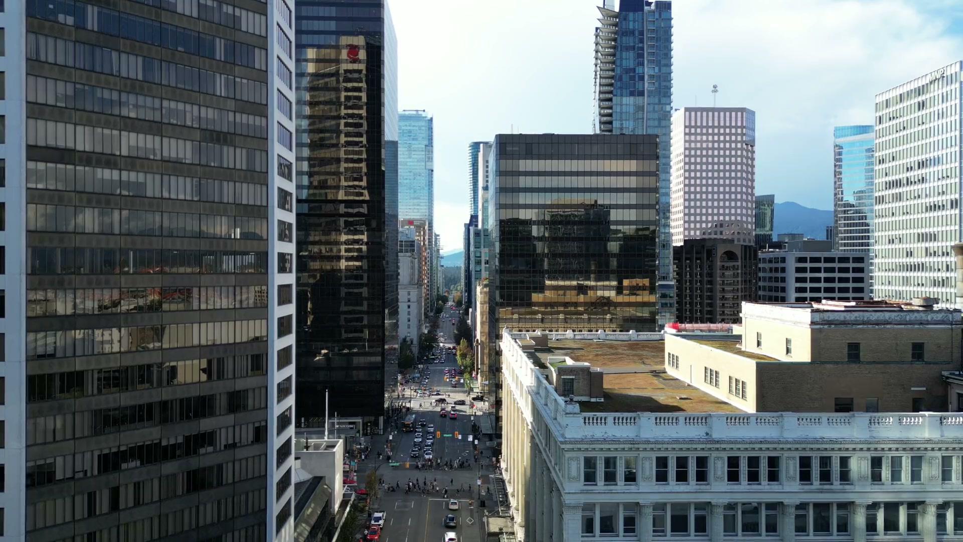 Drifting high among the towers of downtown Vancouver, looking along West Georgia Street across Granville: a crowd in the crosswalk, a terracotta balustrade below, mountains at the street's far end