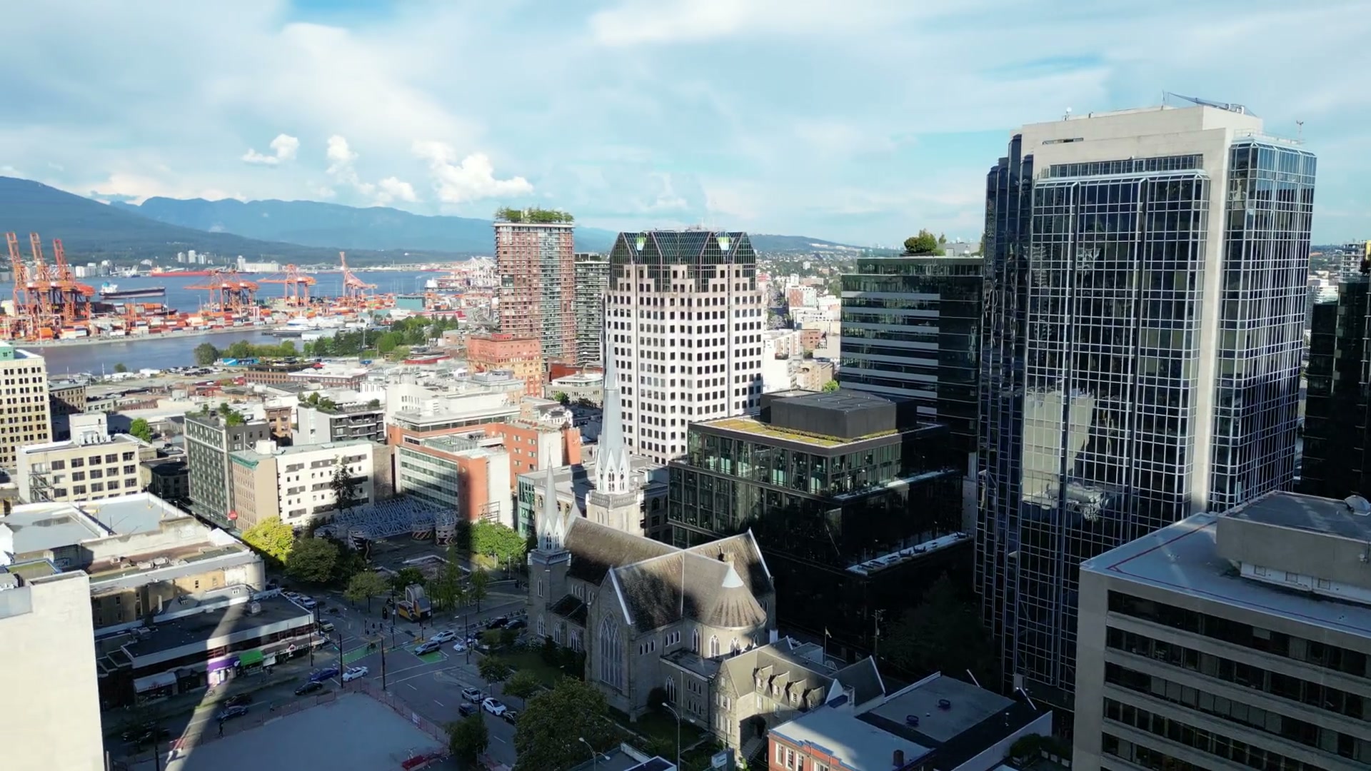 Along West Georgia Street past a terracotta block and a London Drugs sign, then turning and rising over the rooftops until Holy Rosary Cathedral's steeple, the harbour's cranes and the North Shore mountains appear