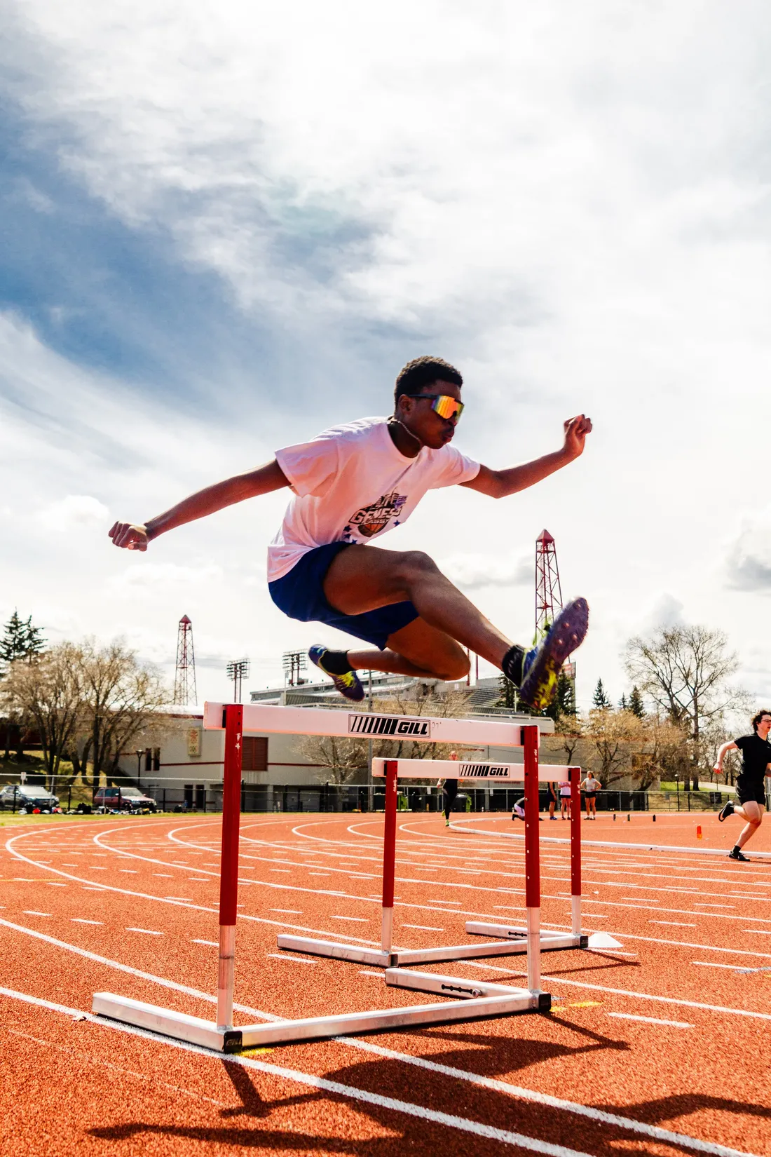 A hurdler mid-flight over a hurdle on an orange outdoor track under a bright cloudy sky