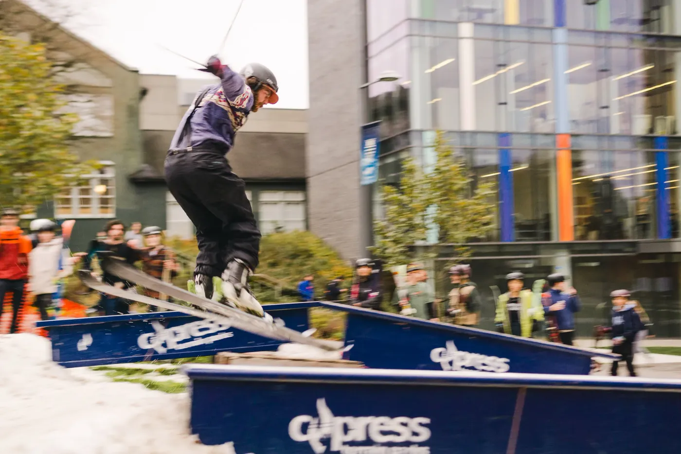 A skateboarder in a helmet dropping off a metal rail at an outdoor plaza skate park while onlookers watch