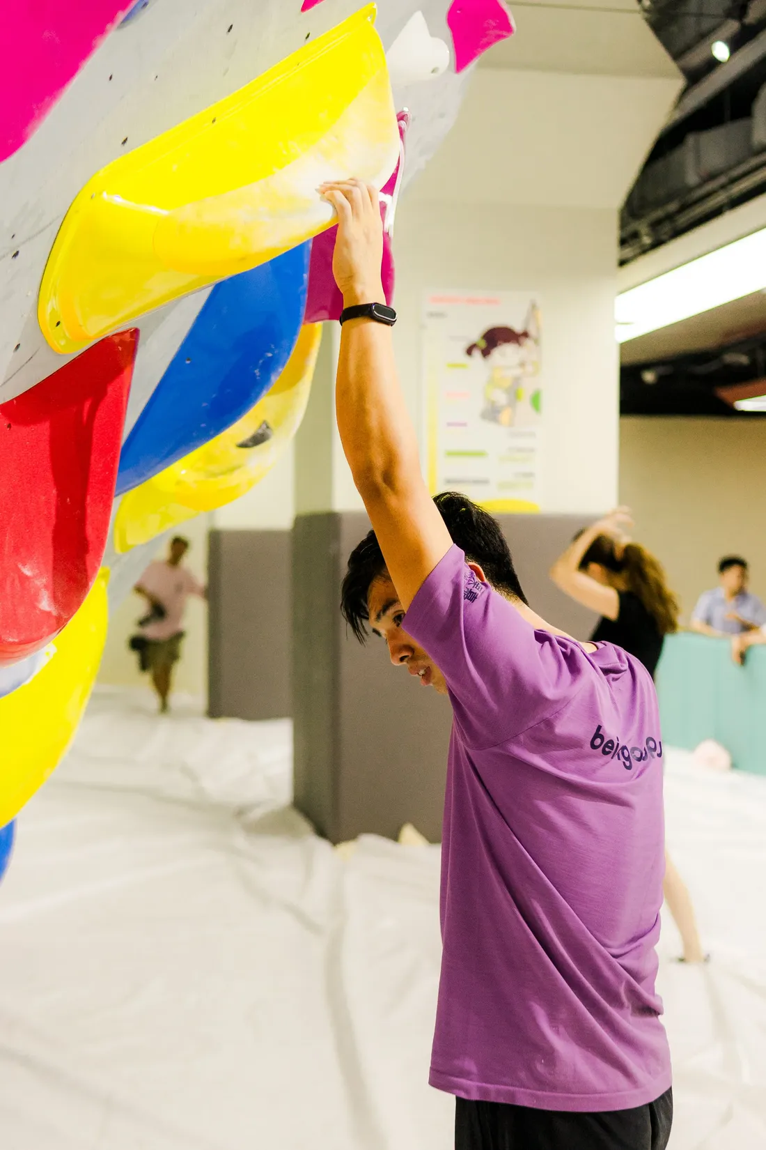 A climber in a purple shirt reaching for a yellow hold on a brightly coloured indoor climbing wall