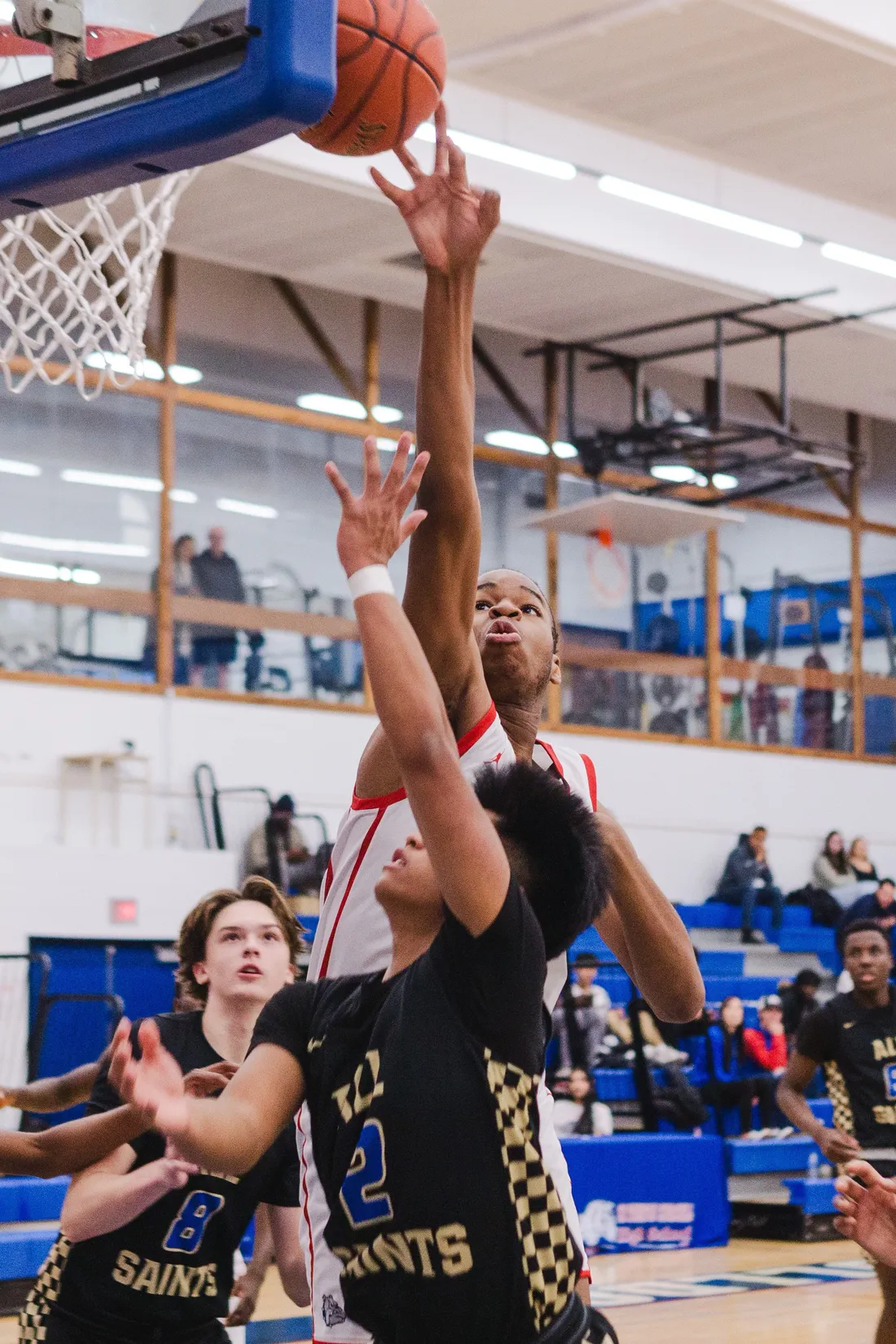 Three high-school basketball players stretching for a rebound under the hoop as a crowd watches from the bleachers