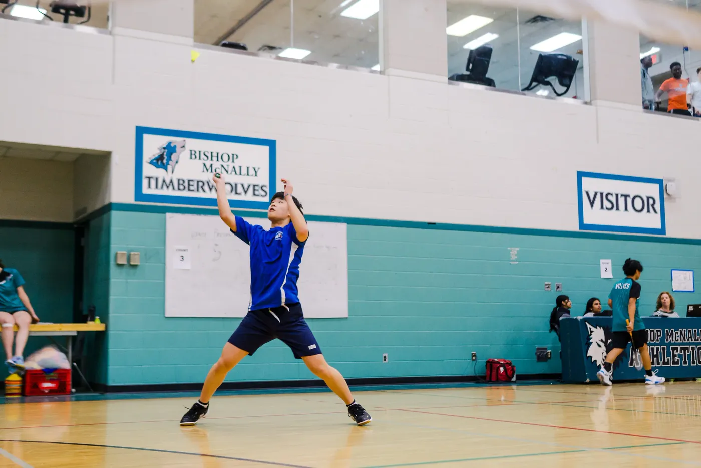 A badminton player winding into a serve in a teal-walled school gym