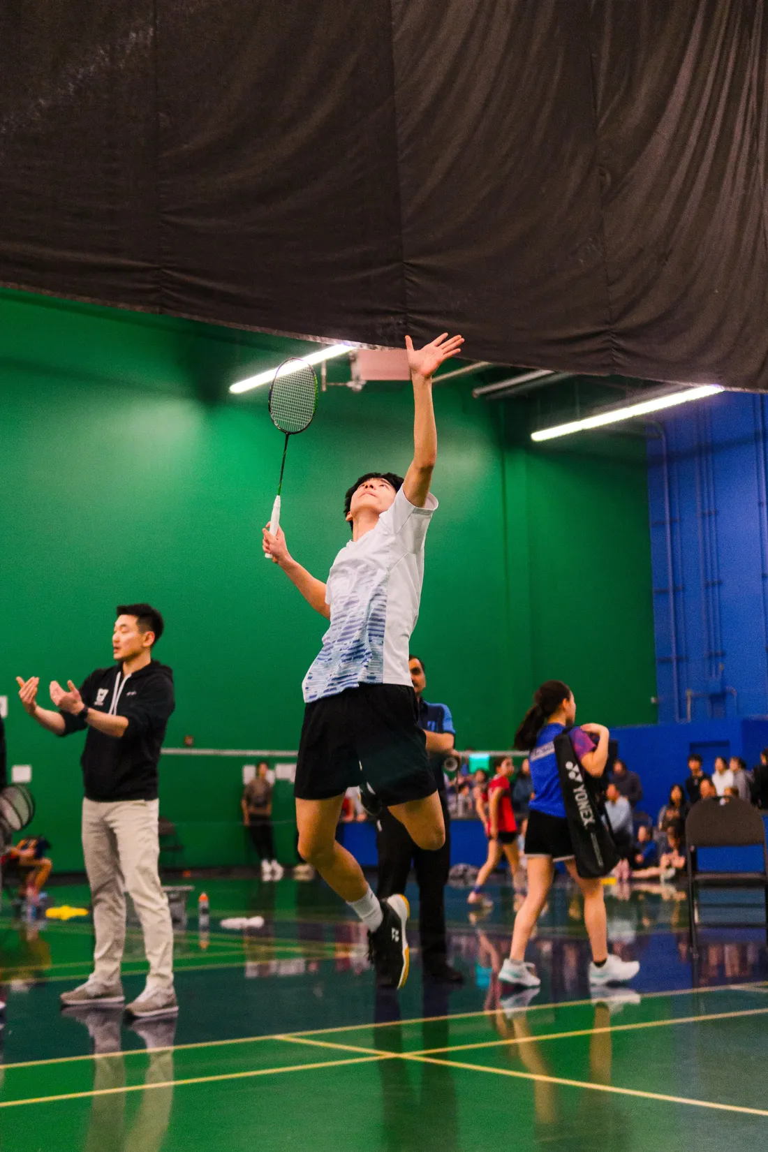 A badminton player leaping into an overhead smash on a green indoor court, teammates watching from the sideline