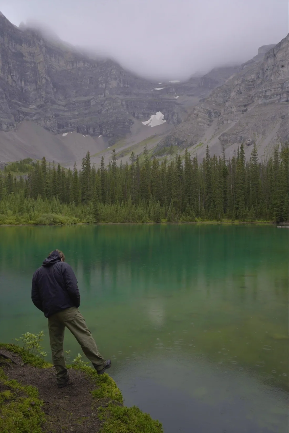 A figure at the edge of an emerald cirque lake in rain, cliffs rising into cloud