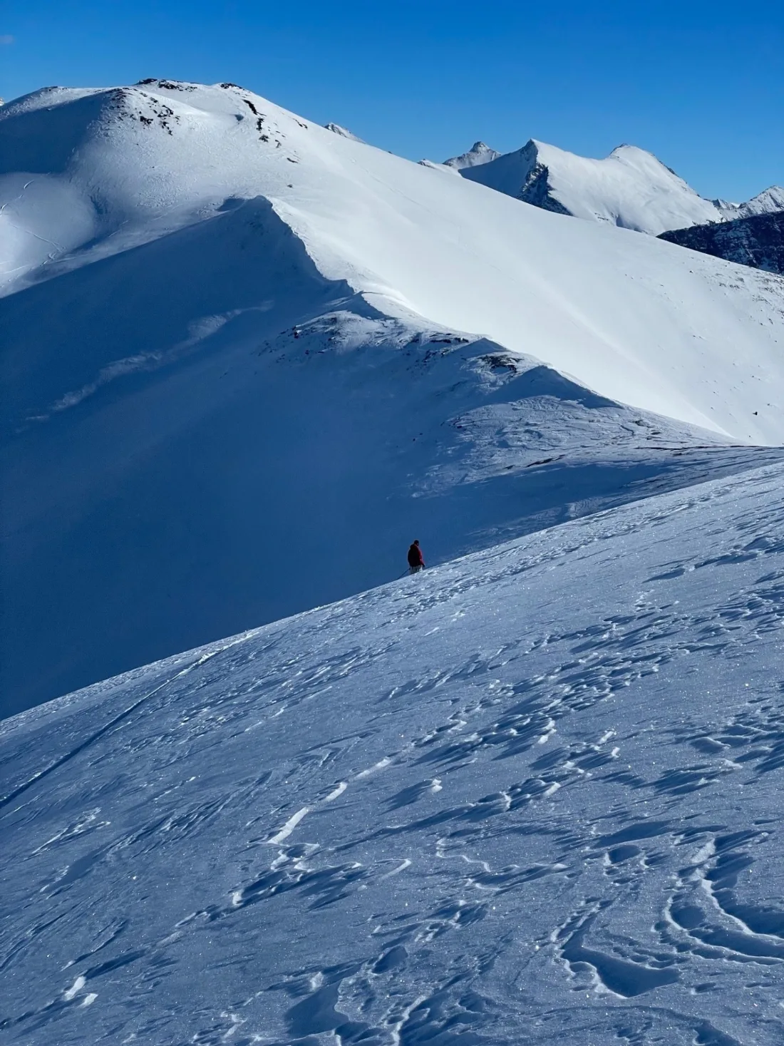 A lone skier on a wind-carved ridge, cornice curling over the drop, high peaks behind