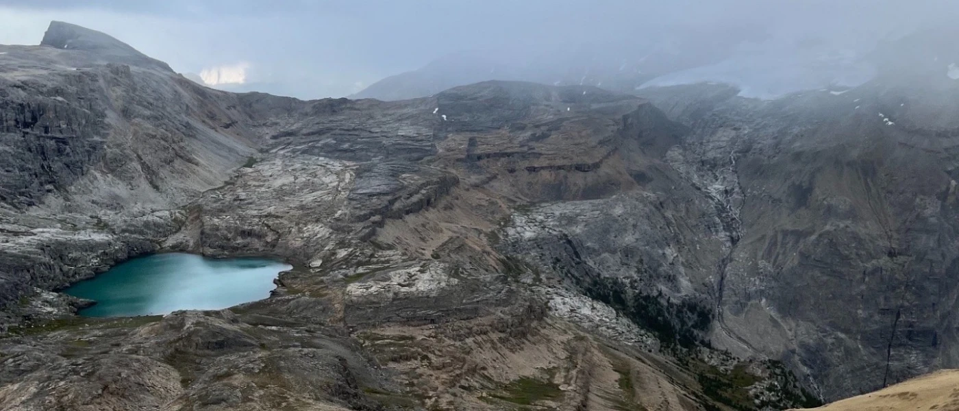 A lone hiker crossing a wide ochre scree slope high above a turquoise tarn, grey limestone benches and a glacier lost in storm cloud beyond