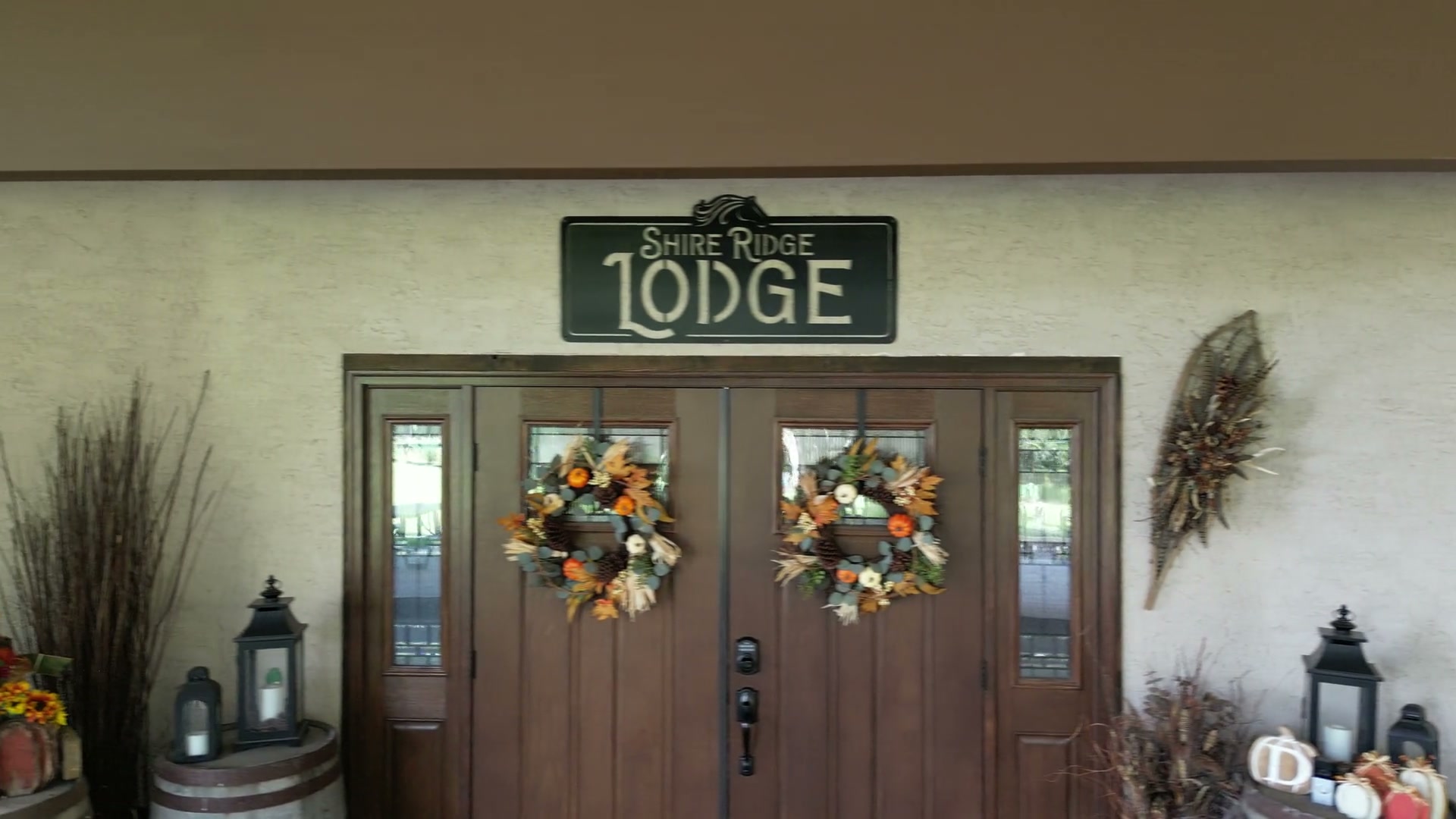 Sliding across the front of Shire Ridge Lodge near Bragg Creek: a stucco wall, the Shire Ridge Lodge sign above double wooden doors, autumn wreaths, lanterns, a wooden barrel, pumpkins and potted flowers