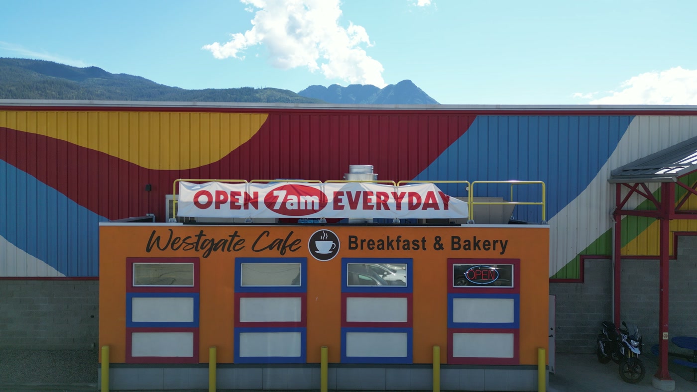 Westgate Cafe held head-on in Salmon Arm: OPEN 7am EVERYDAY on the rooftop banner, the name and the painted cup on the orange front, four framed windows, a motorcycle by the market door