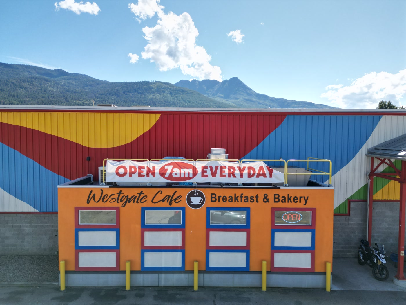 Westgate Cafe head-on in Salmon Arm: its name in black script, a painted coffee cup, Breakfast & Bakery, four windows framed in red and blue, a lit OPEN sign, and the rooftop banner