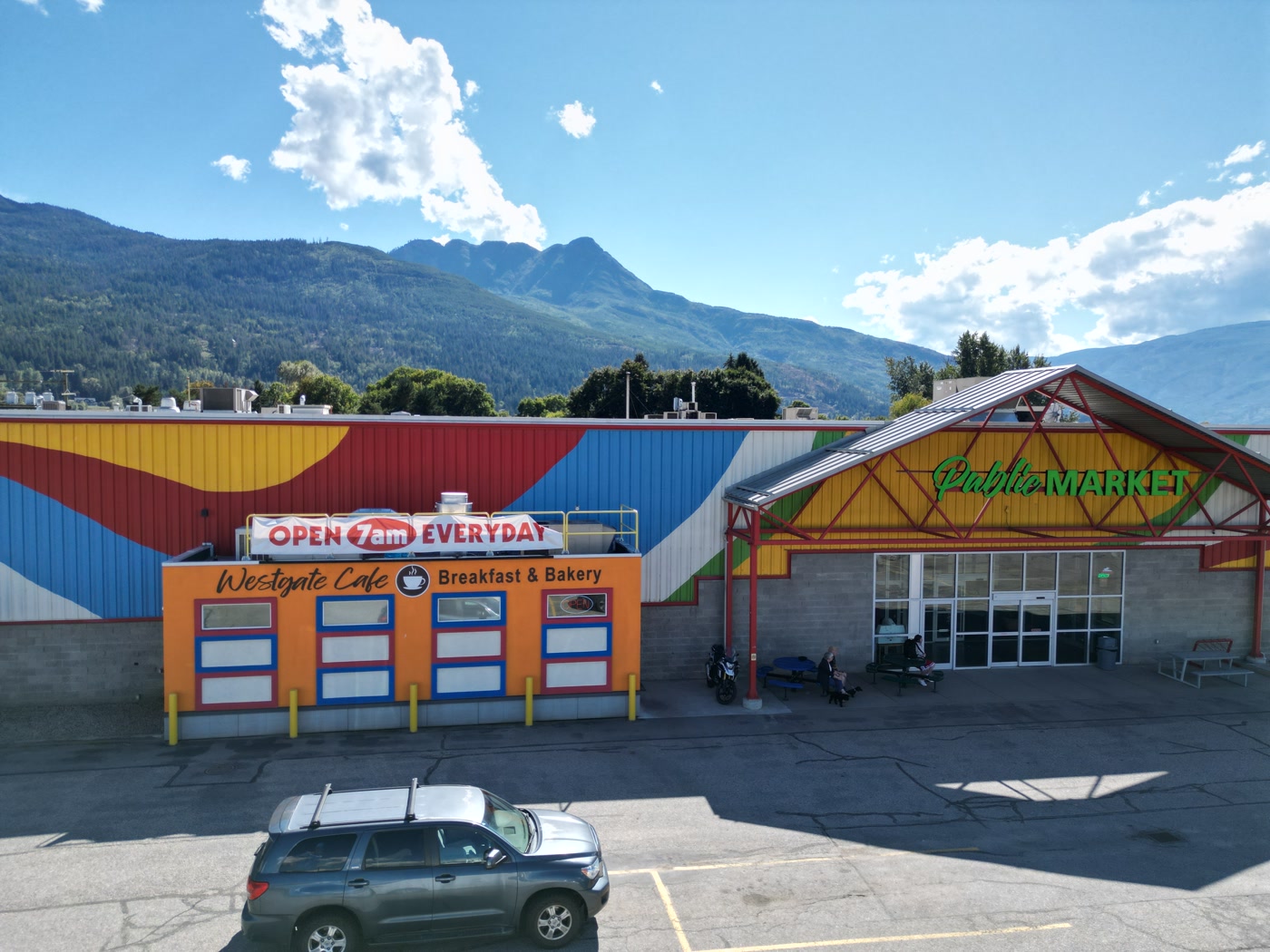 The orange front of Westgate Cafe beside the red steel gable and green Public Market lettering in Salmon Arm, a grey SUV in the lot, and Mount Ida's peak above the painted wall