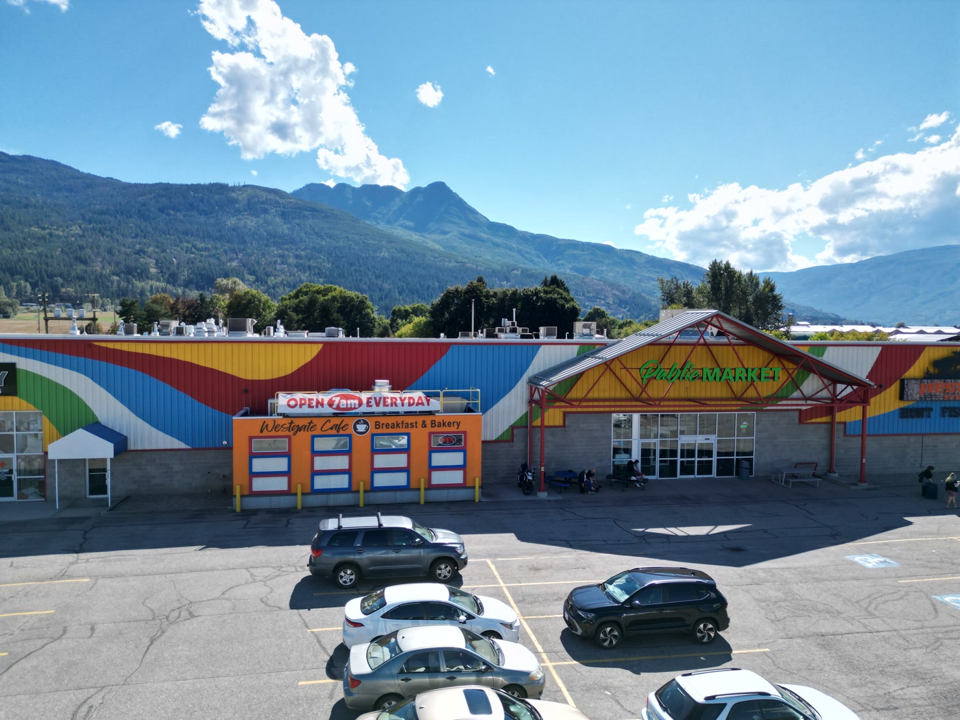 The painted front of Westgate Public Market in Salmon Arm from above the lot: the orange café, the red steel gable, a row of parked cars, the trees, and Mount Ida under cloud