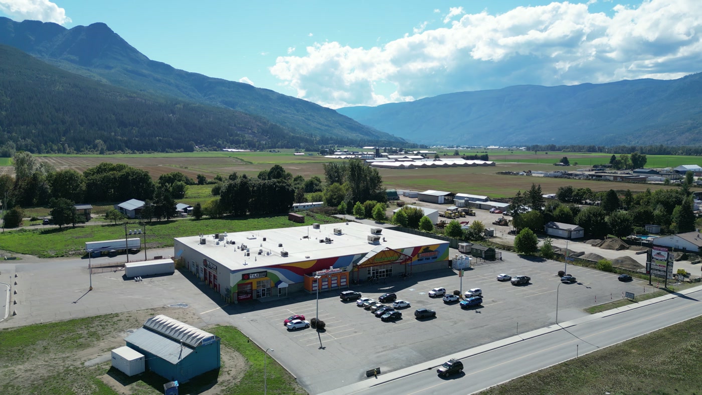 Westgate Public Market small in the valley at Salmon Arm from high above: the white roof and full lot, a teal hut, fields and trees, a road, and Mount Ida's crags on the left