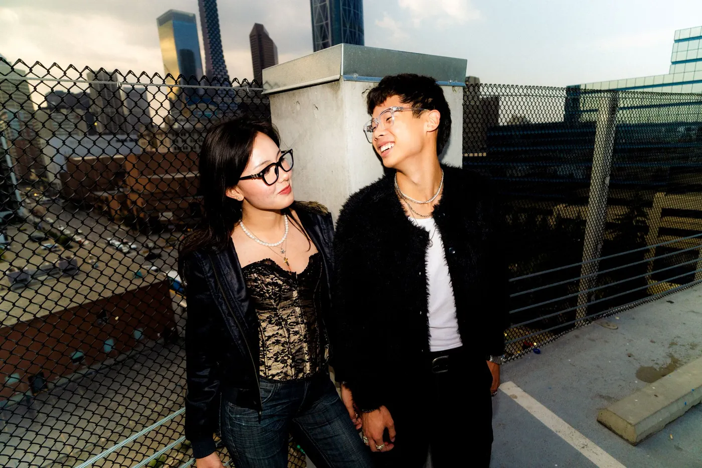 A couple in leather jackets standing hand in hand against a chain-link fence and industrial yard