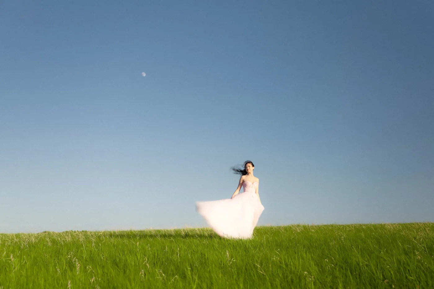 A woman in a white dress standing small in a vast green field under a clear evening sky