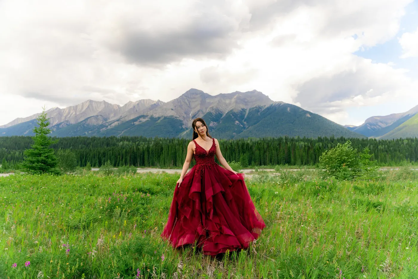 A woman in a deep red ruffled gown standing in a green meadow below a mountain range
