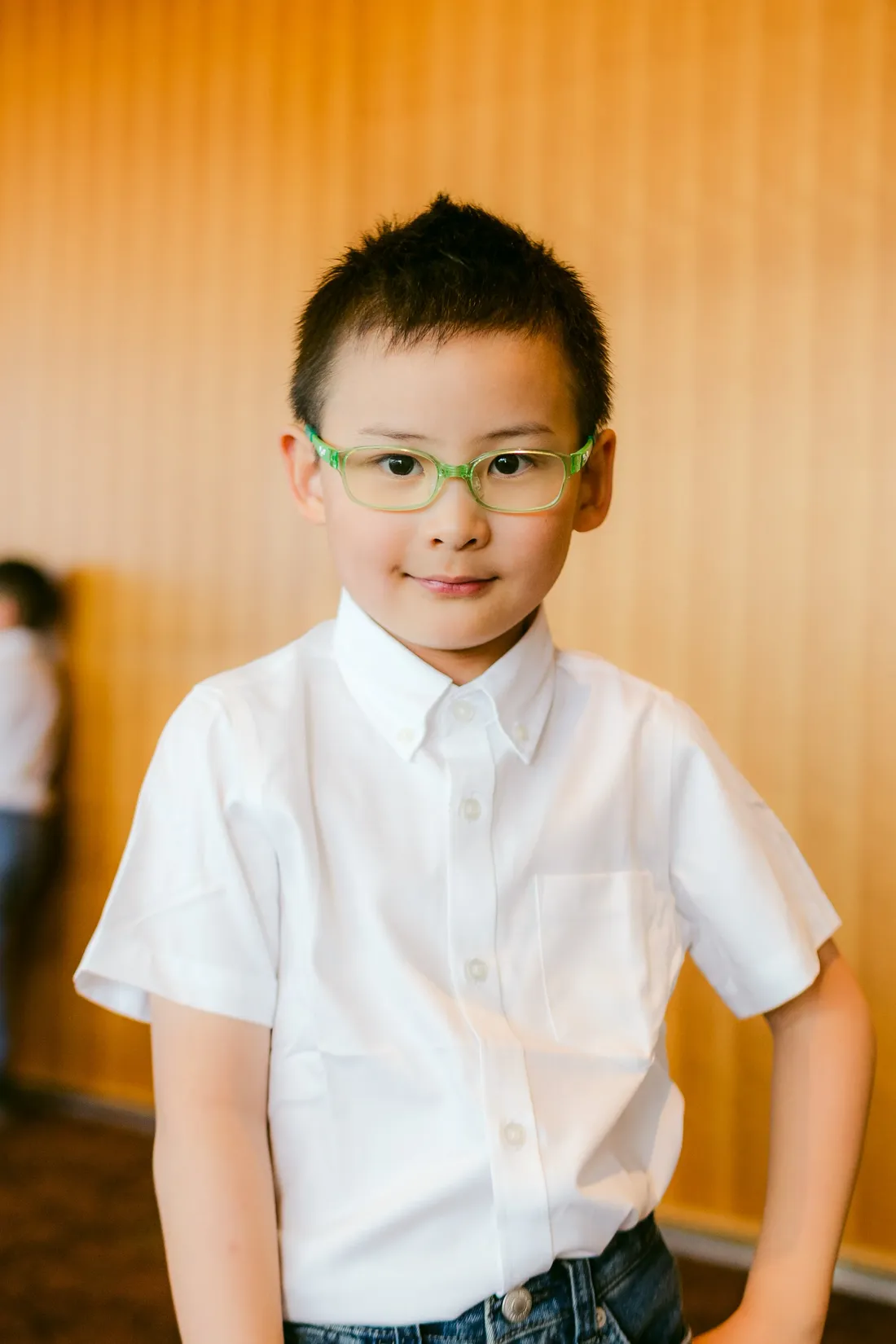 A boy in a white shirt and green glasses photographed against a warm wooden backdrop