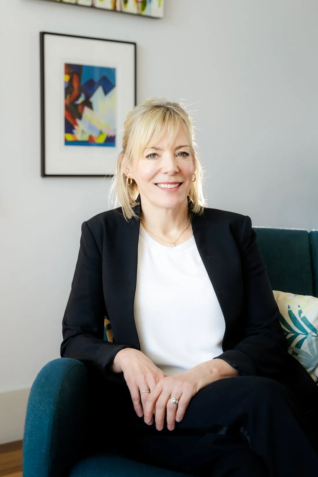 A woman in a black blazer seated on a teal sofa, framed art on the wall behind her