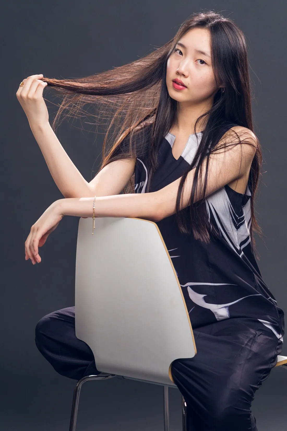 A studio portrait of a woman seated backwards on a white chair against a grey backdrop