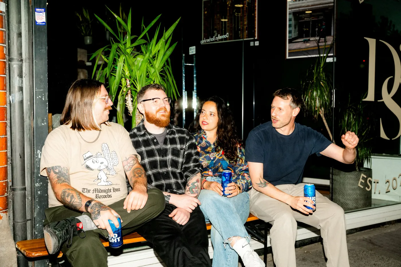 Four people sitting with drinks on a bench outside a dark storefront