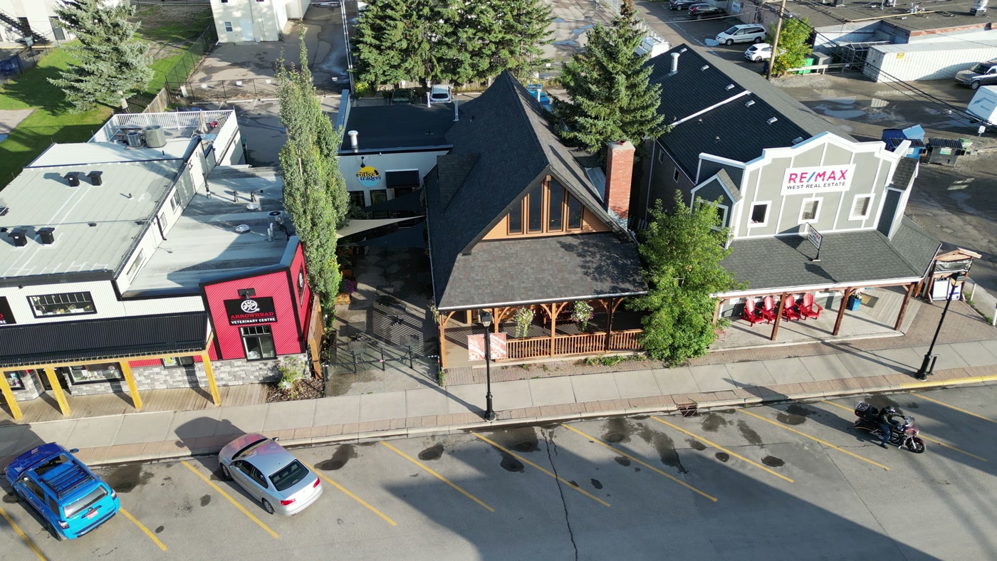 Cochrane Coffee Traders on 2nd Avenue West from above: the timber gable and porch between a red veterinary clinic and a grey real-estate office, a motorcyclist passing, parked cars and long shadows