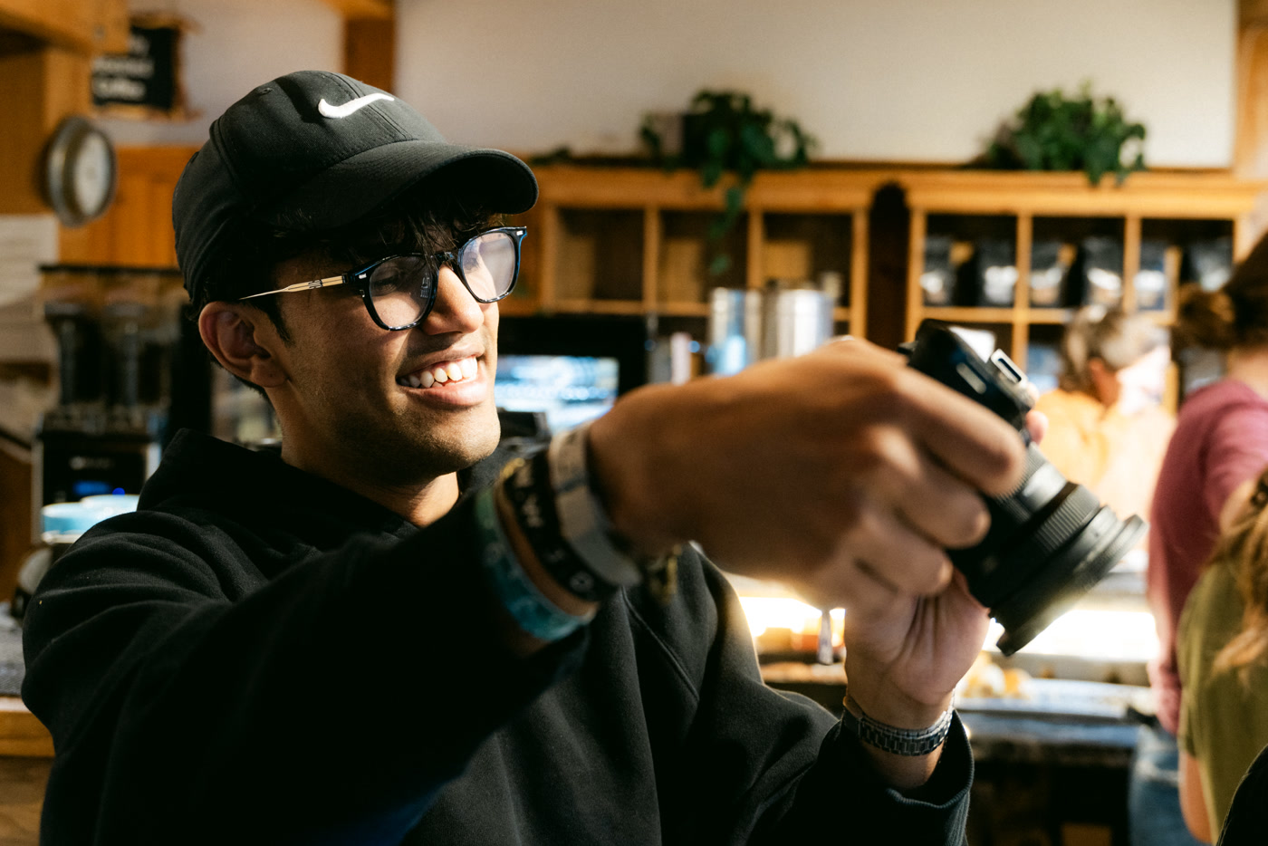 A man in a black cap and glasses smiling as he holds out a camera inside Cochrane Coffee Traders, a round wall clock, timber shelving and potted plants soft behind him