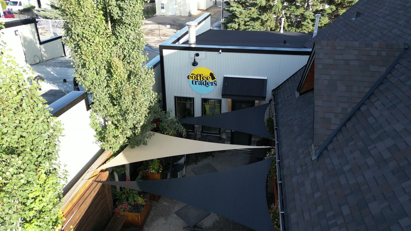 The round yellow-and-blue coffee traders sign in the courtyard of Cochrane Coffee Traders, a coffee pot tipped into its top, on a white corrugated-metal wall under a gooseneck lamp, above charcoal and cream shade sails