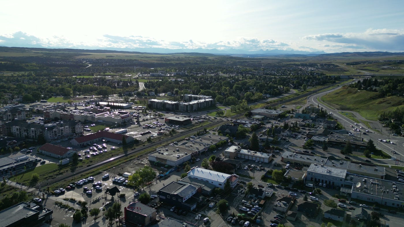 Downtown Cochrane from high above: flat roofs and parking lots, apartment blocks, a rail line running across town, a highway curving past a green hillside and under an overpass, and a pale line of mountains