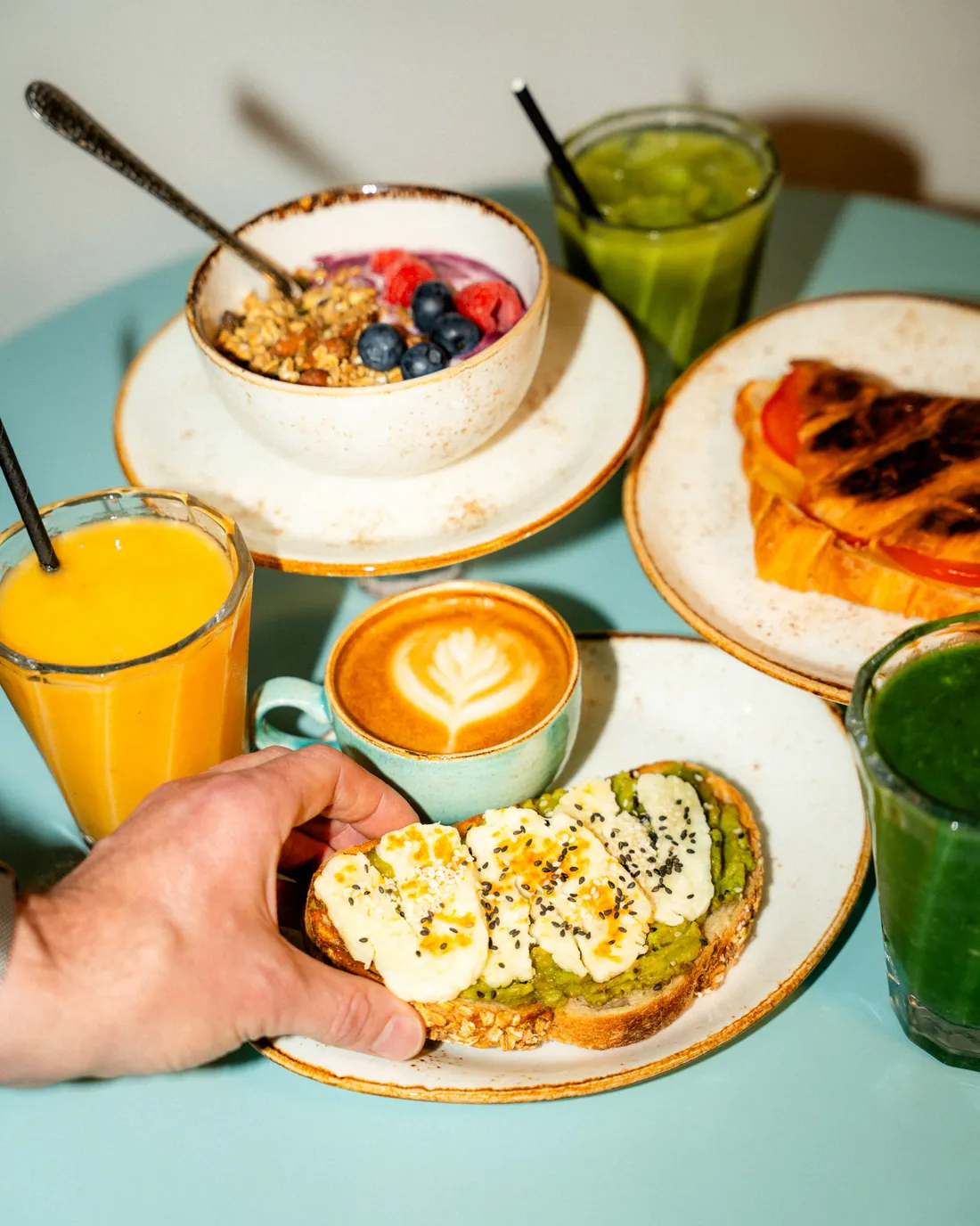 A hand lifting avocado toast from a table set with a latte, orange juice, granola bowl and green juice