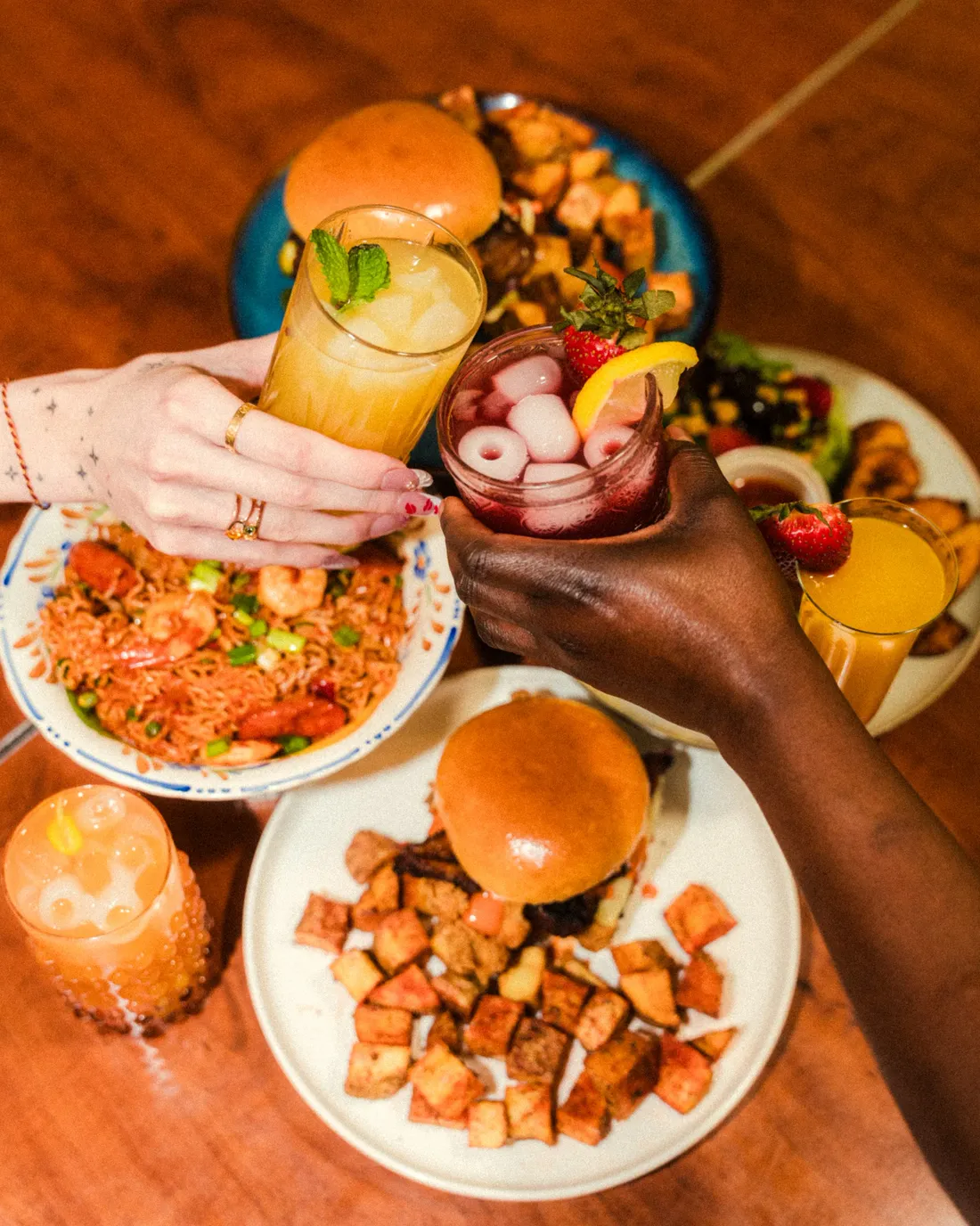 Hands reaching across a brunch table of burgers, rice, potatoes and cocktails
