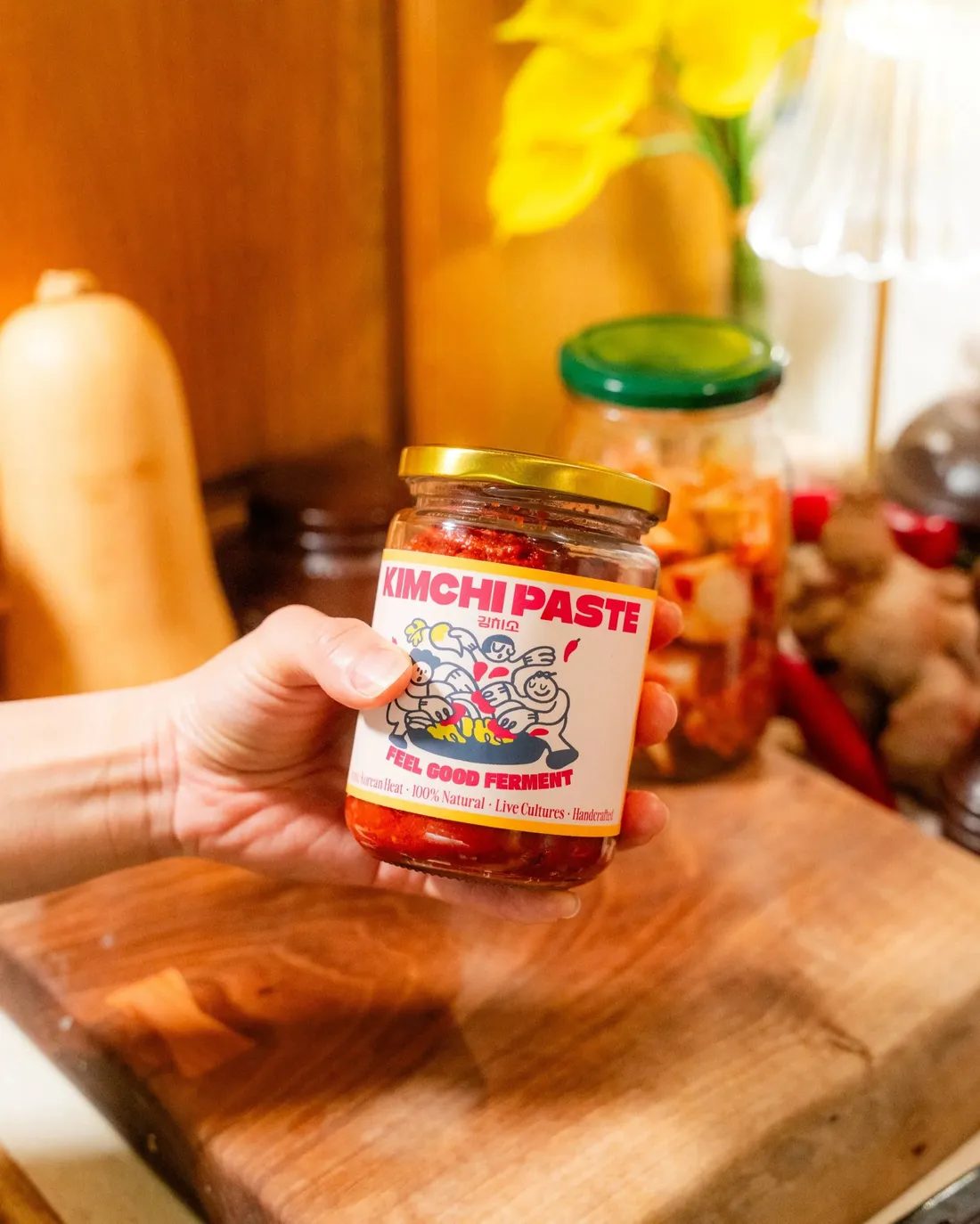 A hand holding a jar of kimchi paste above a wooden board of ginger, chillies and fermented vegetables