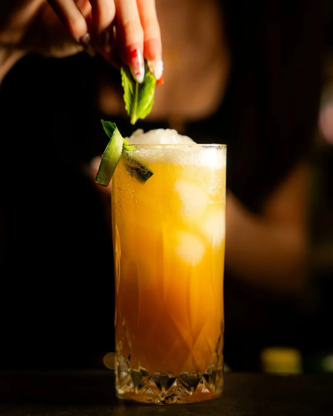 A tall layered cocktail being garnished with a leaf in a dark bar