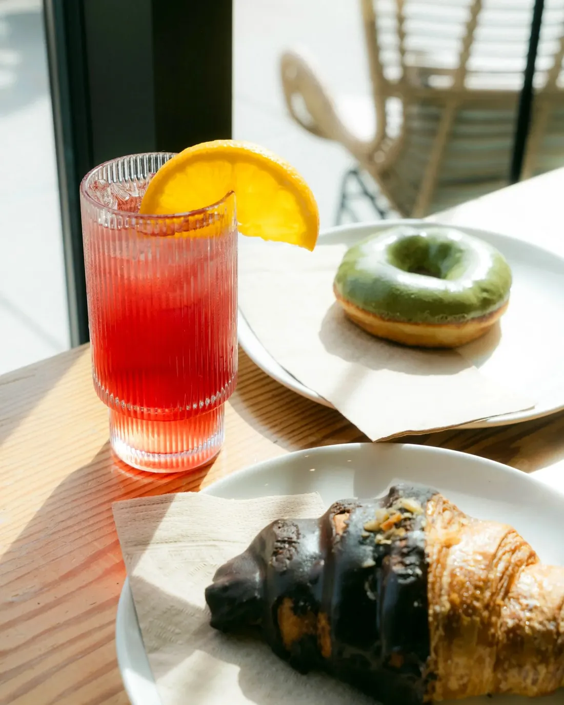 An iced red drink with an orange slice beside a matcha-glazed donut and a chocolate croissant on a sunlit table