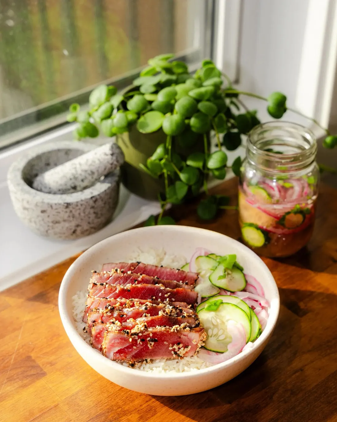 A bowl of seared sesame-crusted tuna with cucumber and pickled onion on a wood table in window light
