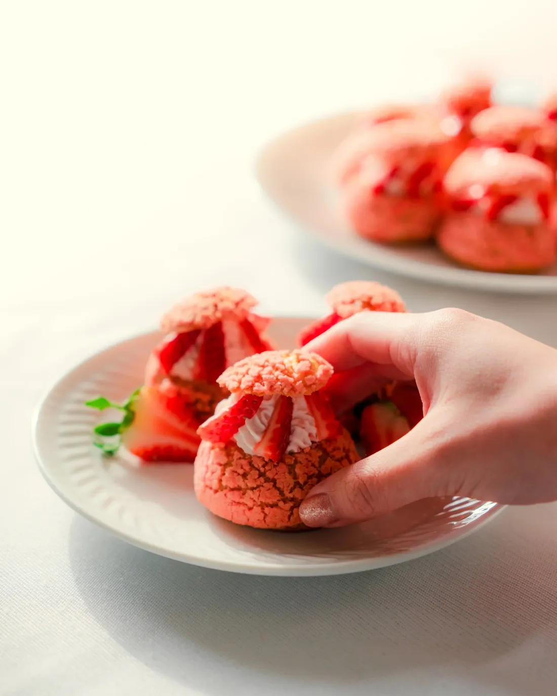 A hand lifting a strawberry-topped cookie from a plate in soft pale light